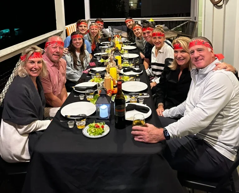 A large group of people enjoying a hibachi dinner at a long, elegantly set table on a cozy outdoor porch at night. Everyone is smiling, wearing matching red bandanas, and surrounded by plates of food, drinks, and decorative lighting.