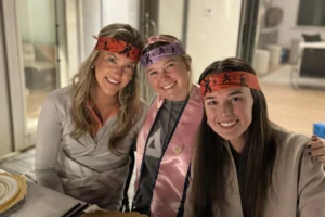 Three women enjoying a lively hibachi dinner party at Alys Beach, seated at a beautifully set table with gold-rimmed plates and fresh salad, celebrating with festive headbands