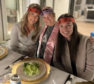 Three women enjoying a lively hibachi dinner party at Alys Beach, seated at a beautifully set table with gold-rimmed plates and fresh salad, celebrating with festive headbands